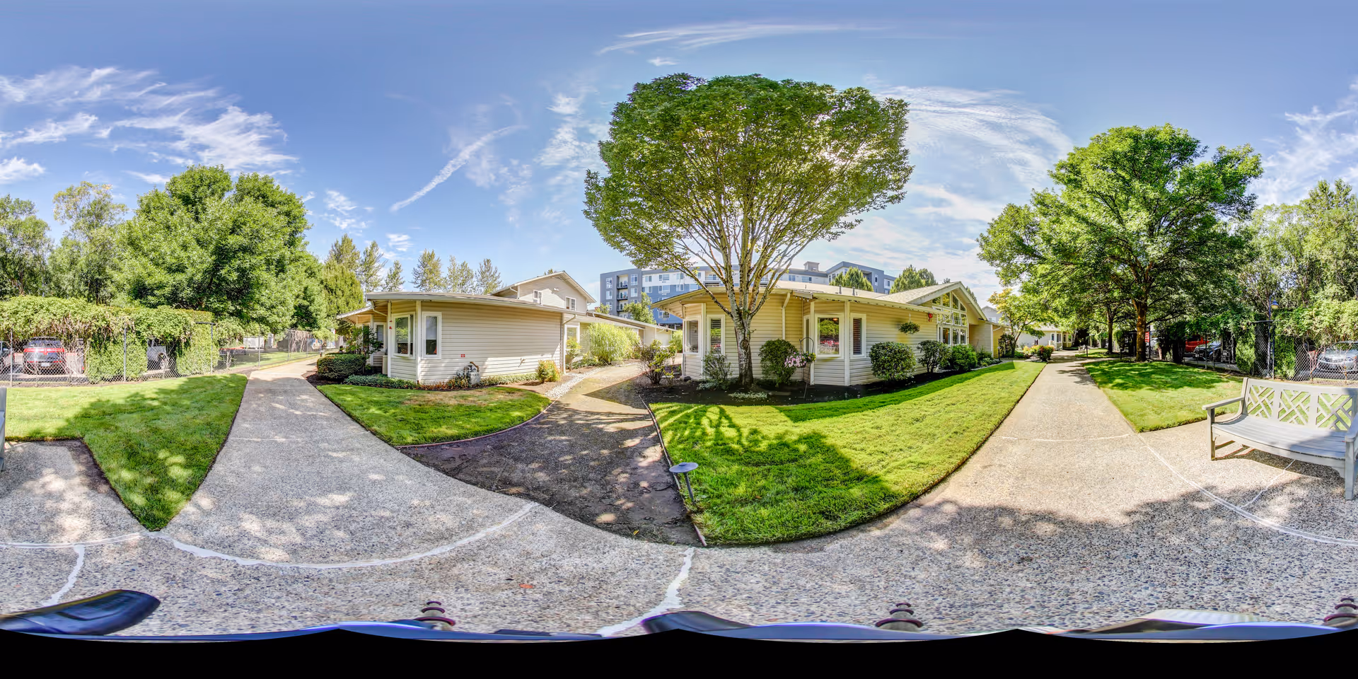 Outdoor view of a senior living facility with paved walkways, green lawns, trees, and single-story buildings under a partly cloudy sky.