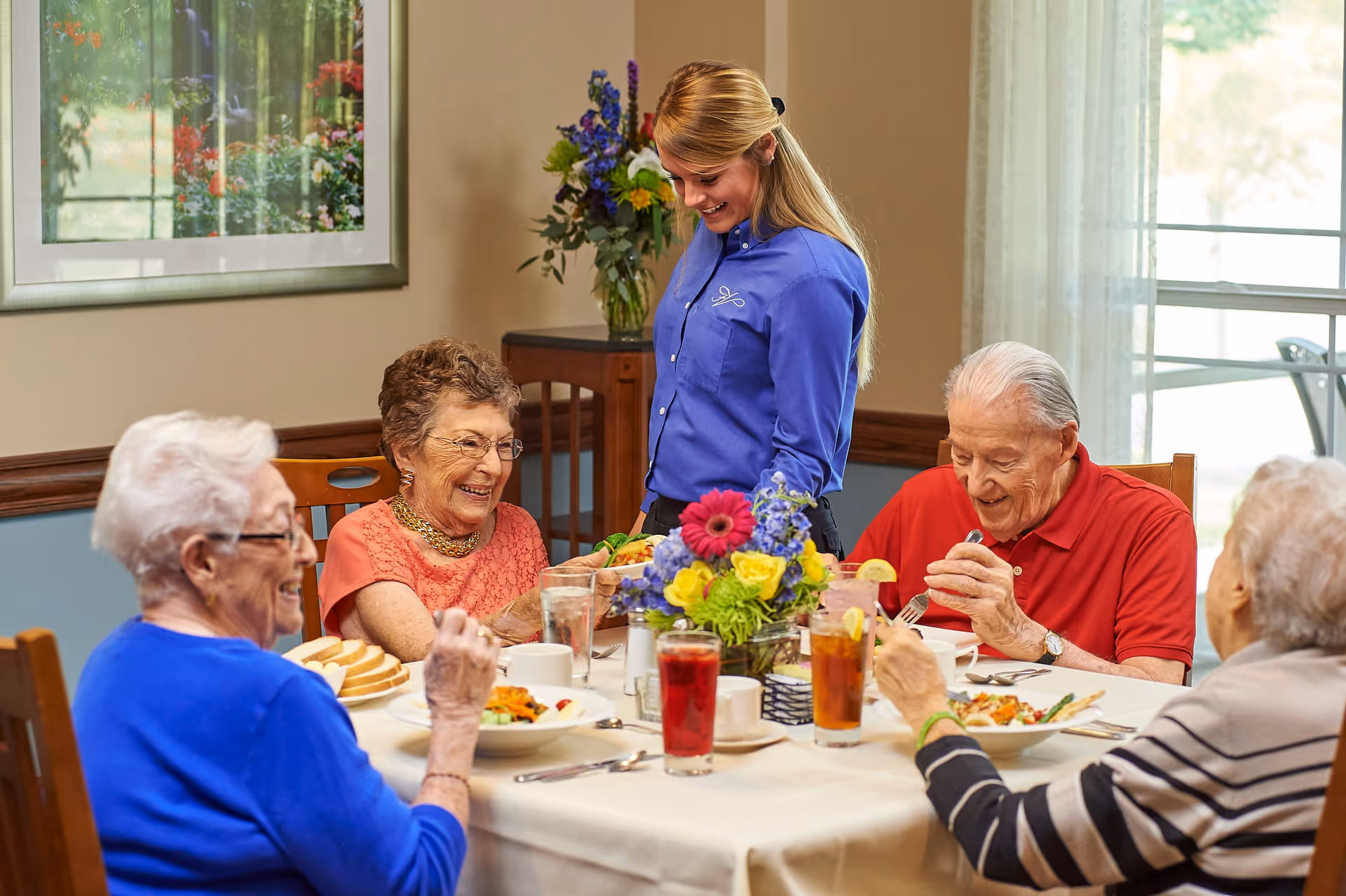 A group of elderly people sitting around a dining table enjoying a meal together, with a smiling staff member standing and serving them. The table is set with plates of food, drinks, and a colorful flower centerpiece. The room has a window with sheer curtains and a framed picture on the wall.