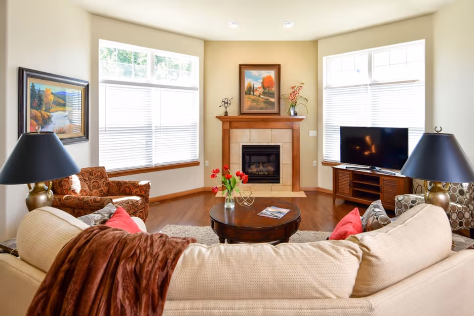 Well-lit living room with a beige sofa facing a fireplace, round coffee table, TV, and large windows.