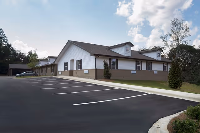 Exterior view of a single-story senior living facility building with white siding and brown trim, surrounded by a parking lot with marked spaces and some trees in the background under a partly cloudy sky.