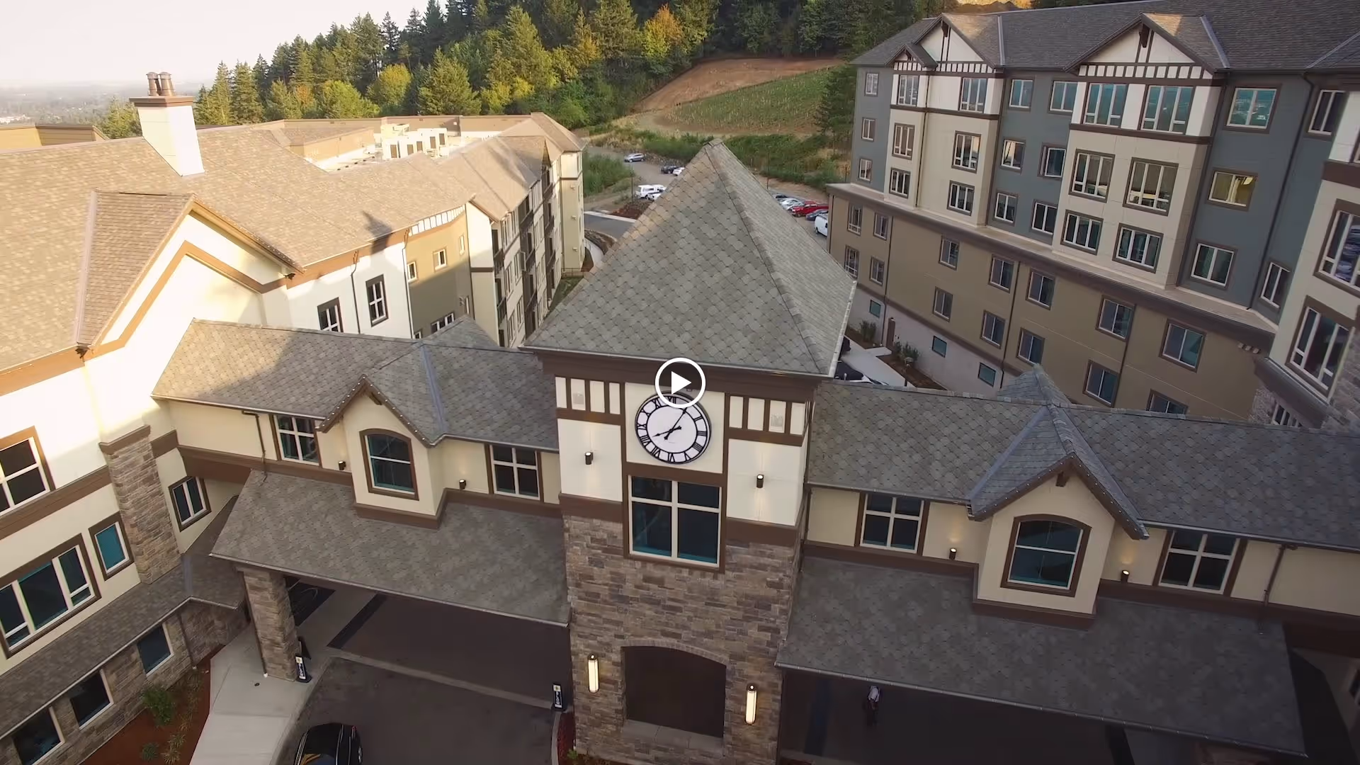 Aerial view of the Touchmark in the West Hills senior living facility showing a large building complex with a central clock tower, multiple windows, and a covered entrance. The surrounding area includes trees and a parking lot with several cars.