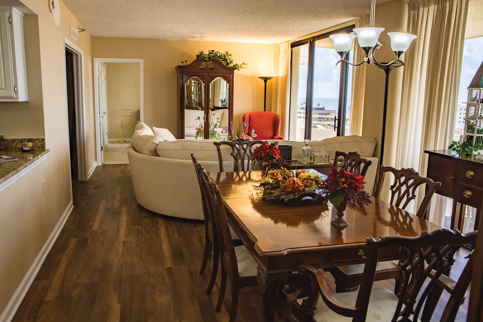 Interior view of a senior living facility showing a dining area with a wooden table and six chairs, decorated with floral centerpieces. Beyond the dining area is a living room with a curved beige sofa, a red armchair, a wooden cabinet with mirrors, and large windows with curtains letting in natural light.