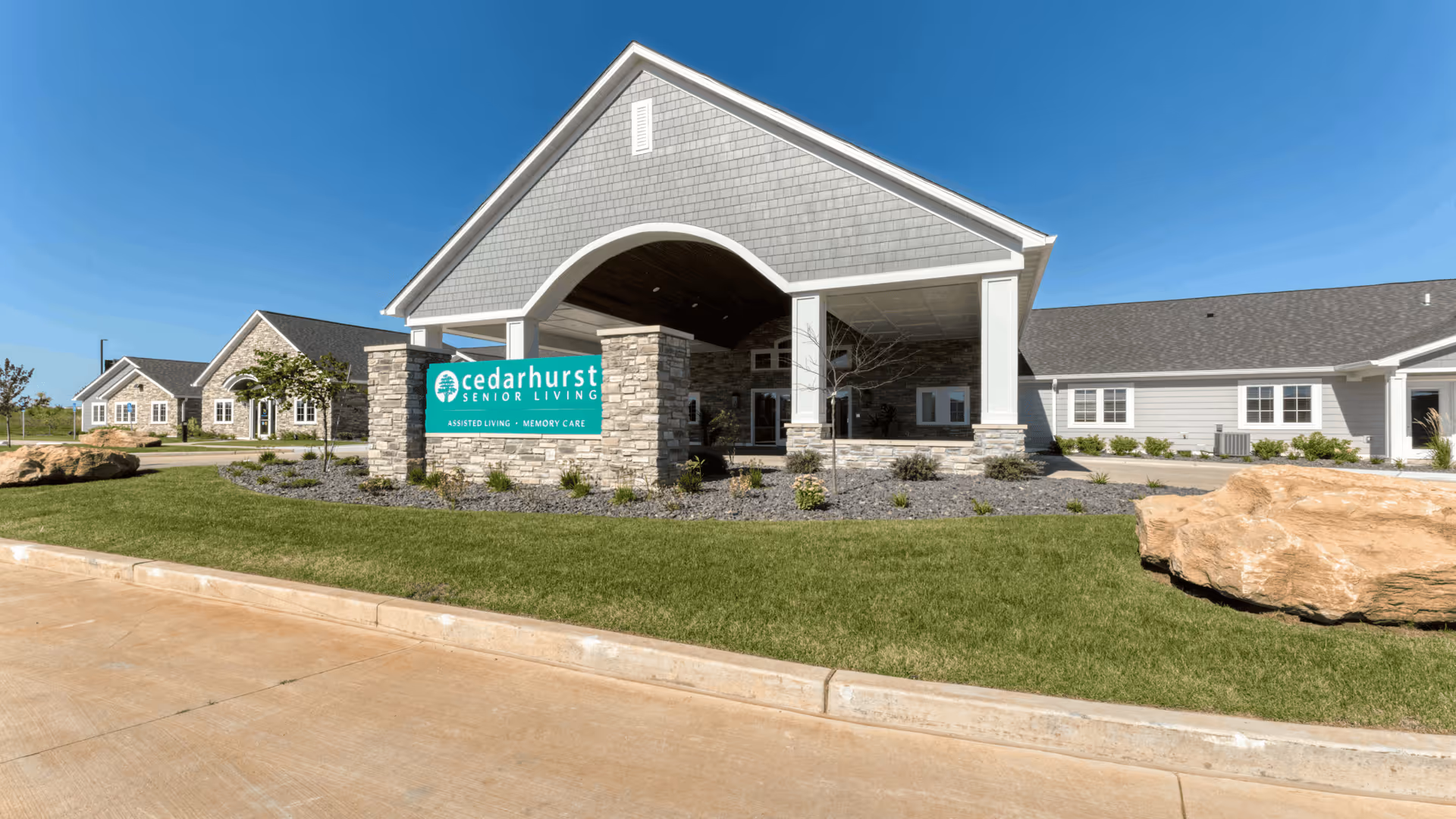 Front exterior of Cedarhurst Senior Living showing the covered entrance, stone sign, and landscaped lawn under a clear blue sky.