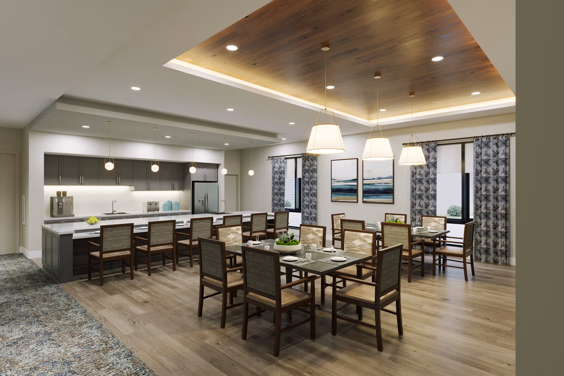 A spacious dining area in a senior living facility featuring multiple tables with chairs arranged for group seating. The room has a modern kitchen with a long island and bar stools, pendant lighting, and large windows with patterned curtains. The ceiling has recessed lighting and a wooden accent panel. Two abstract paintings hang on the wall.