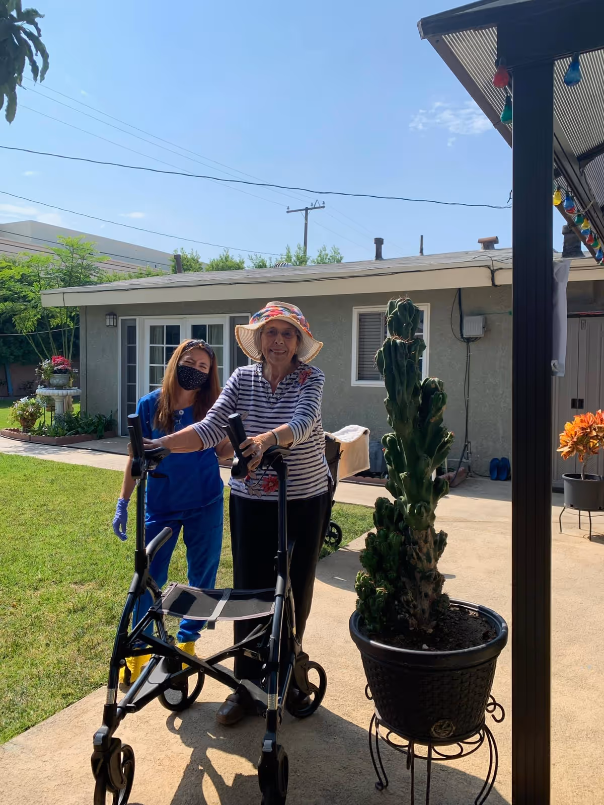 An elderly woman wearing a sun hat and striped shirt is standing outdoors with a walker. A caregiver in blue scrubs, a face mask, and gloves stands beside her. They are in a garden area with a building in the background and a potted cactus plant in the foreground.