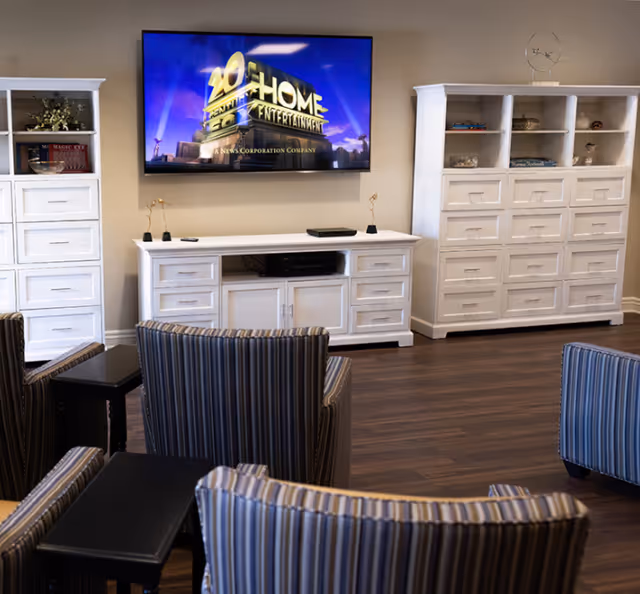 A cozy living room area with a wall-mounted flat-screen TV displaying the 20th Century Fox Home Entertainment logo. The room features white wooden cabinets and shelves on either side of the TV, decorated with small items and books. In front of the TV are several striped upholstered chairs with small black side tables. The floor is dark wood, and the walls are painted a neutral color.