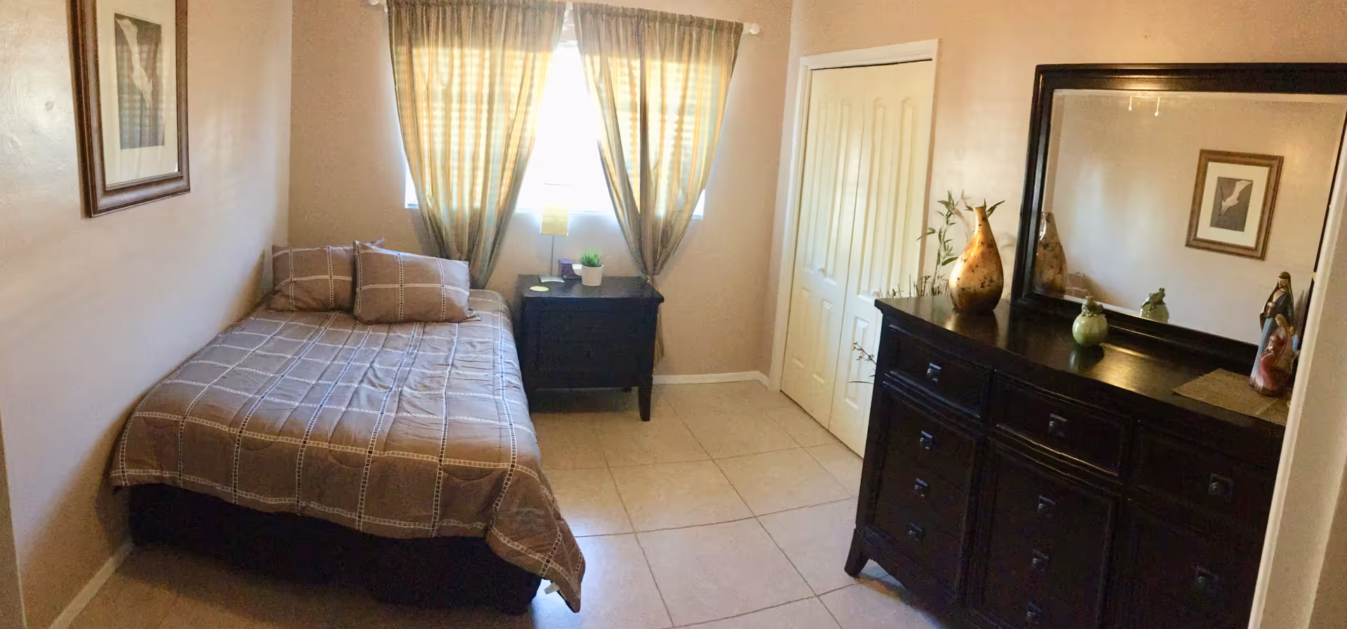 A small bedroom with a bed covered in a brown checkered bedspread and two matching pillows. Next to the bed is a black nightstand with a small lamp and a potted plant. The room has beige walls, a window with beige curtains, a framed picture on the wall, and a large dark wooden dresser with a mirror and decorative items on top.