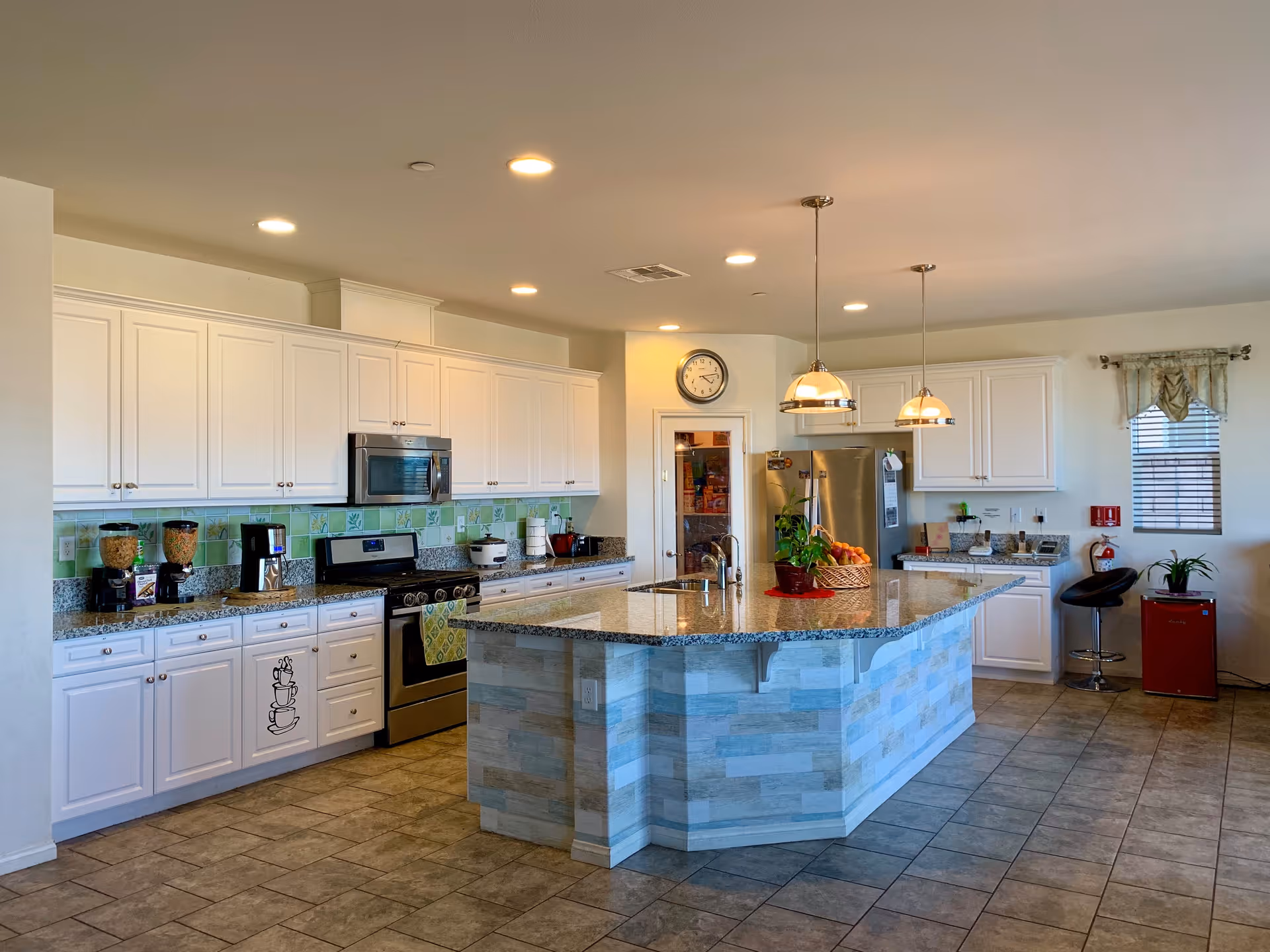 A spacious, modern kitchen with white cabinets, granite countertops, and a large central island with a sink. The kitchen features stainless steel appliances including a stove and refrigerator. There are two pendant lights hanging over the island, a clock on the wall, and a small seating area with a black bar stool and a red mini fridge near a window with blinds and a valance.
