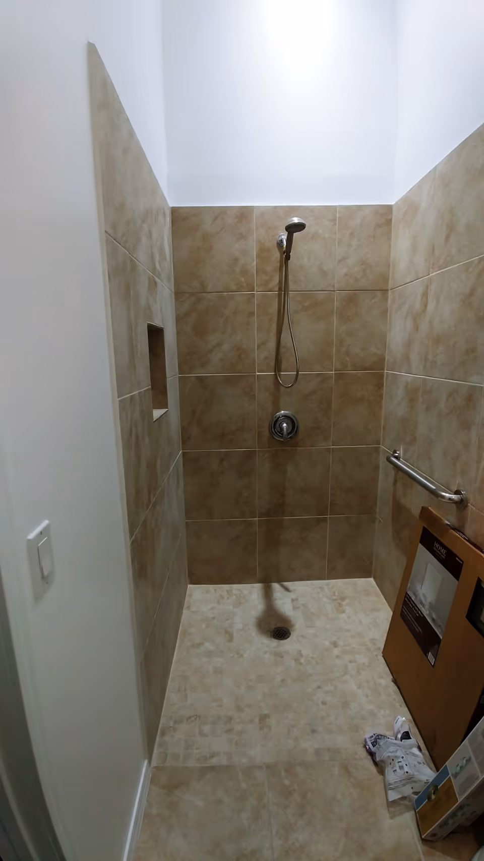 Small tiled walk-in shower with a handheld showerhead, a grab bar, a recessed niche, and cardboard boxes and packaging on the floor.