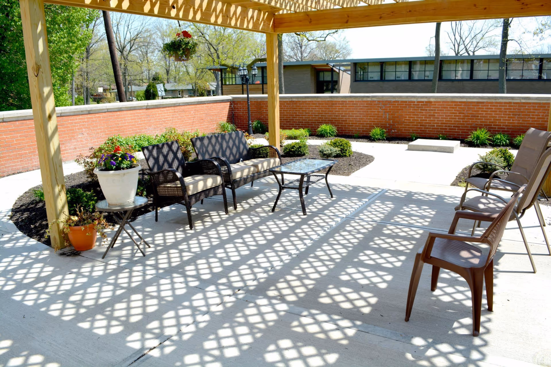 Outdoor patio area with a wooden pergola casting lattice shadows on the concrete floor. The patio has two cushioned wicker chairs, a glass-top table, two brown plastic chairs, and a small round table with potted plants. There is a brick wall enclosing the space and some greenery and trees in the background.