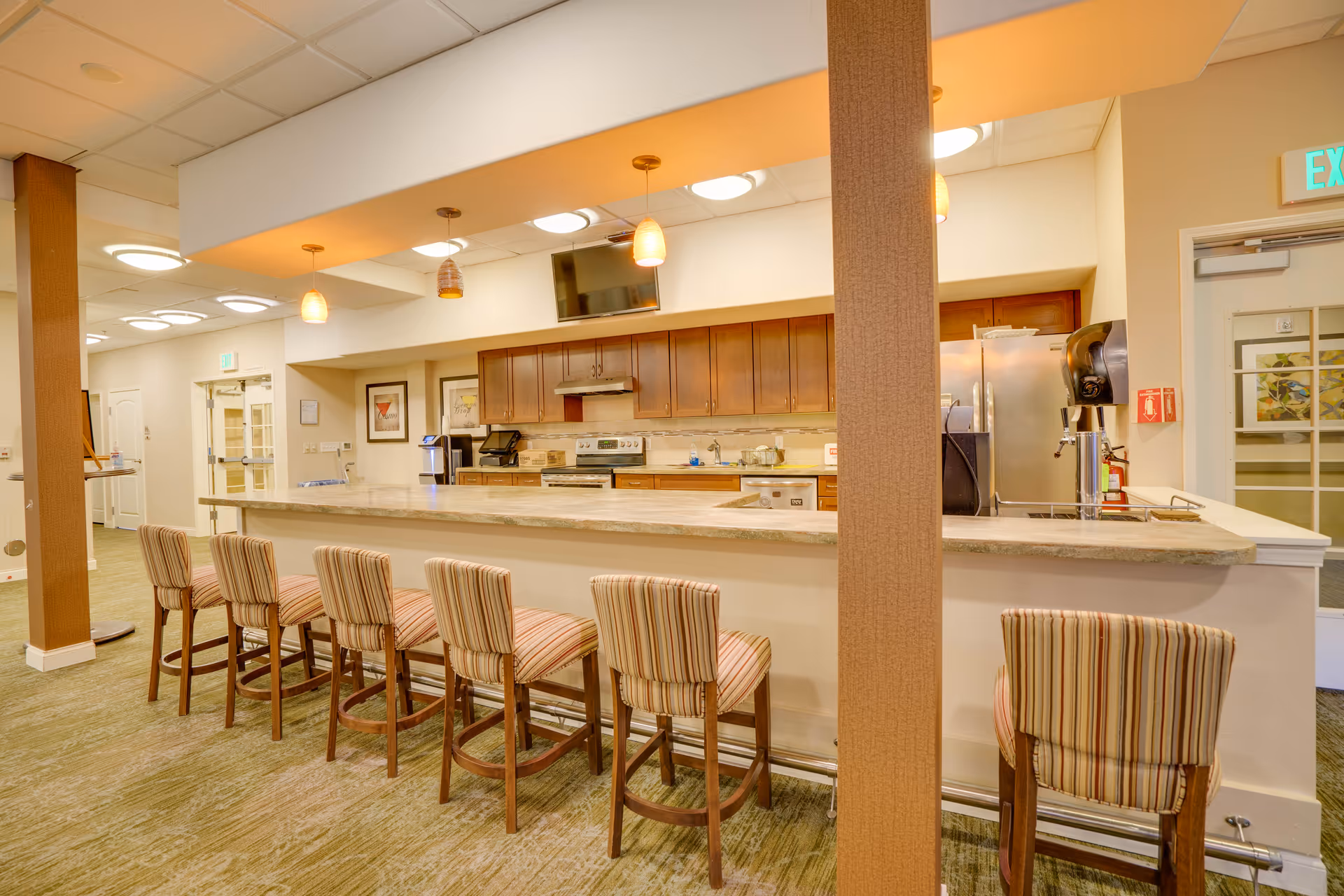 Interior view of a retirement facility kitchen area with a long counter and six striped cushioned bar stools. The kitchen features wooden cabinets, a stove, a refrigerator, a coffee machine, and a mounted TV above the counter. The area is well-lit with ceiling lights and pendant lights hanging over the counter.