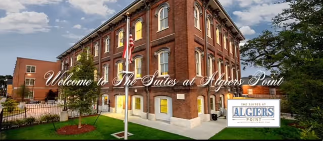 Exterior view of a large, historic brick building with multiple windows, surrounded by a well-maintained lawn and trees. An American flag is displayed on a flagpole in front of the building. The sky is partly cloudy. Text overlay reads 'Welcome to The Suites at Algiers Point' with a logo in the bottom right corner.