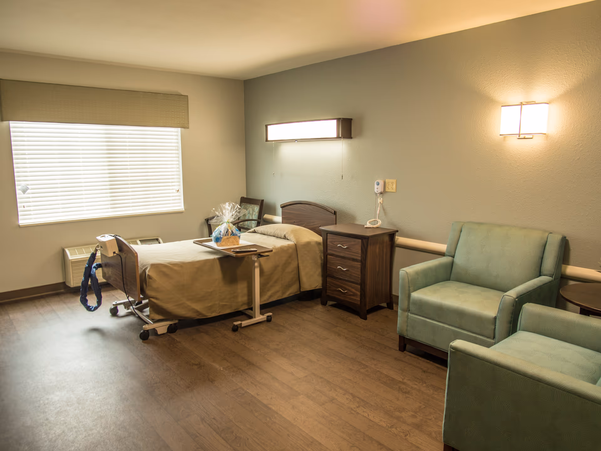 A nursing facility room with a single hospital bed covered in beige bedding, a wooden nightstand with three drawers, a green armchair, and a window with closed blinds. The room has wooden flooring and soft lighting from wall-mounted fixtures.