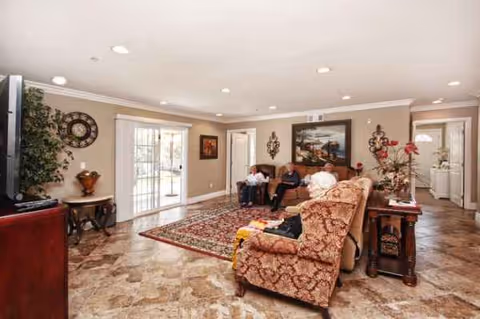 A cozy senior living room with residents seated on patterned furniture and a rug.