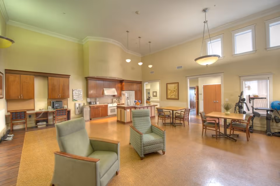 A spacious, well-lit common area featuring a kitchen with wooden cabinets, a refrigerator, stove, and microwave. The room includes several tables with chairs, two comfortable armchairs in the foreground, and exercise equipment near the windows. The walls are painted a light yellow, and there are high windows letting in natural light. Pendant and ceiling lights provide additional illumination.