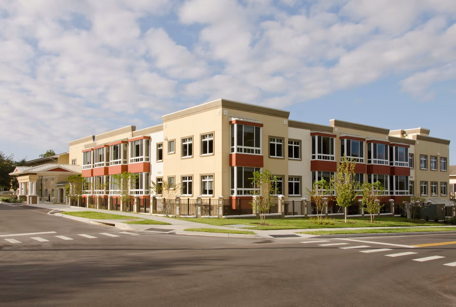 Modern two-story senior living facility building with large windows, landscaped lawn, and a driveway under a partly cloudy sky.