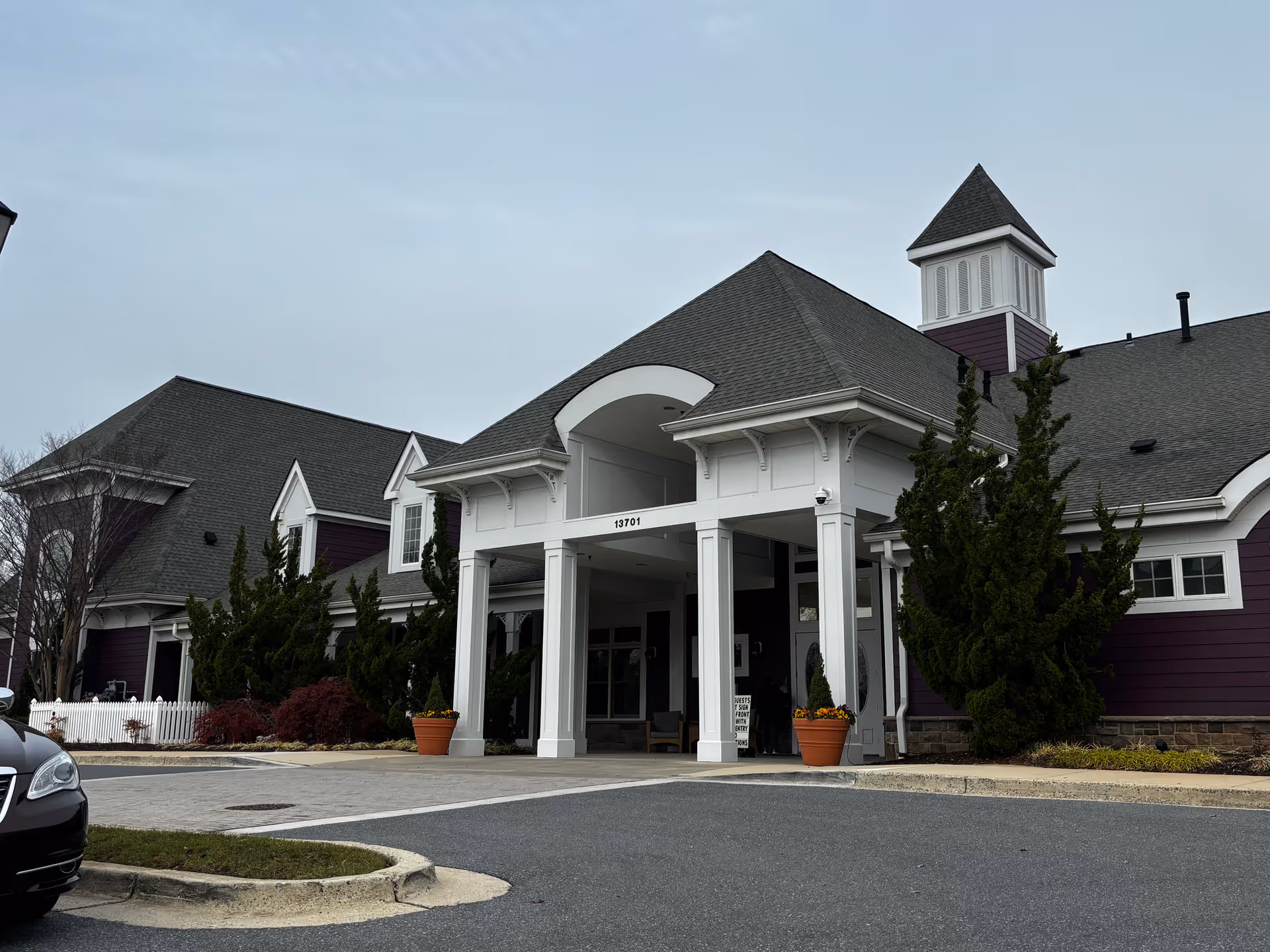 Exterior view of a senior living facility building with a covered entrance supported by white columns. The building has a dark gray roof, purple siding, and white trim. There are two large potted plants with flowers near the entrance and some trees and shrubs along the side. A black car is partially visible on the left side of the image.