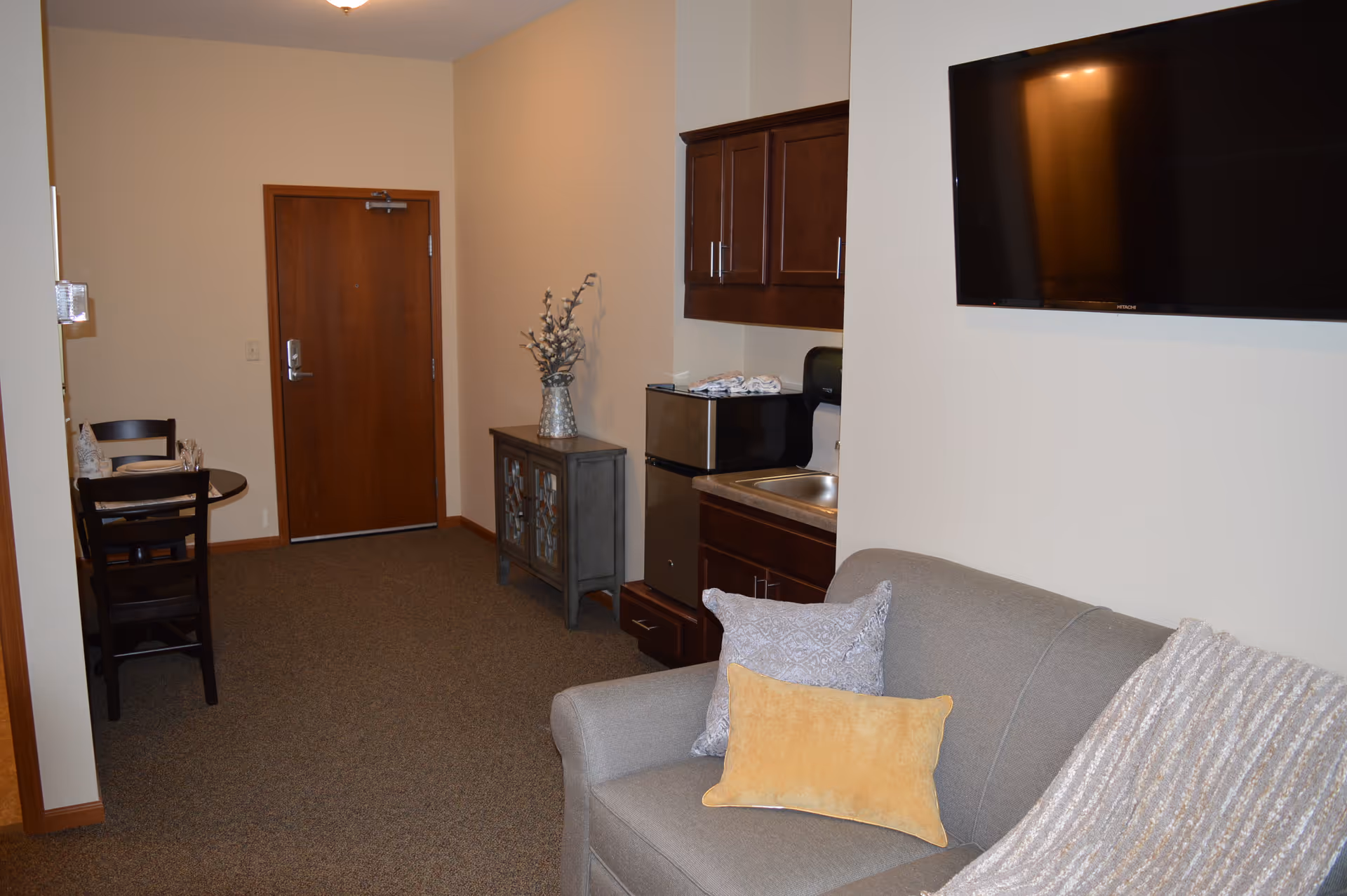 Interior view of a senior living facility room featuring a gray couch with decorative pillows and a throw blanket, a wall-mounted flat-screen TV, a small kitchenette with a sink, microwave, and cabinets, a wooden side table with a vase of flowers, and a small dining table with two chairs near a wooden door.