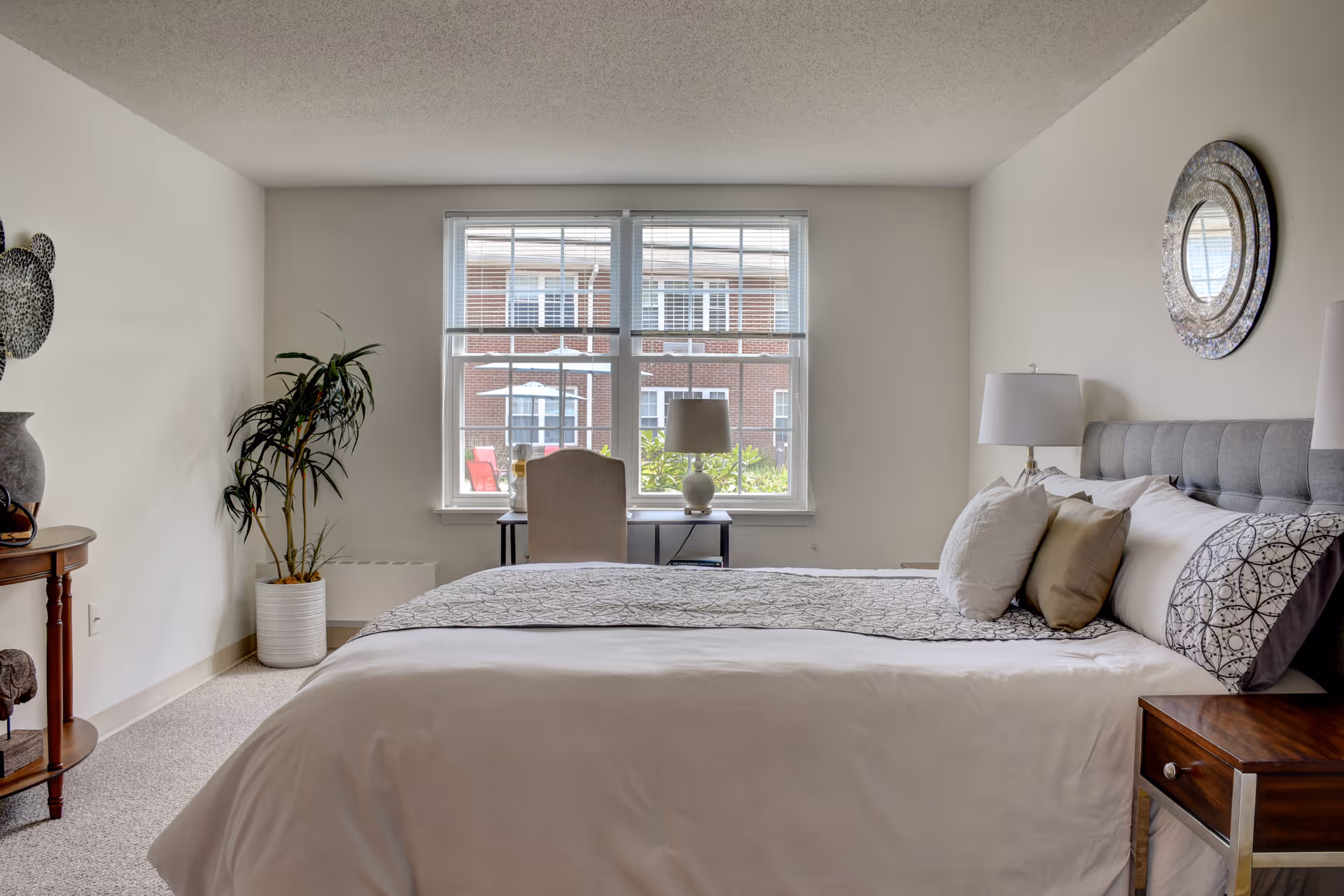 Well-lit bedroom with a neatly made bed, bedside tables and lamps, a desk by a large window, and a potted plant.