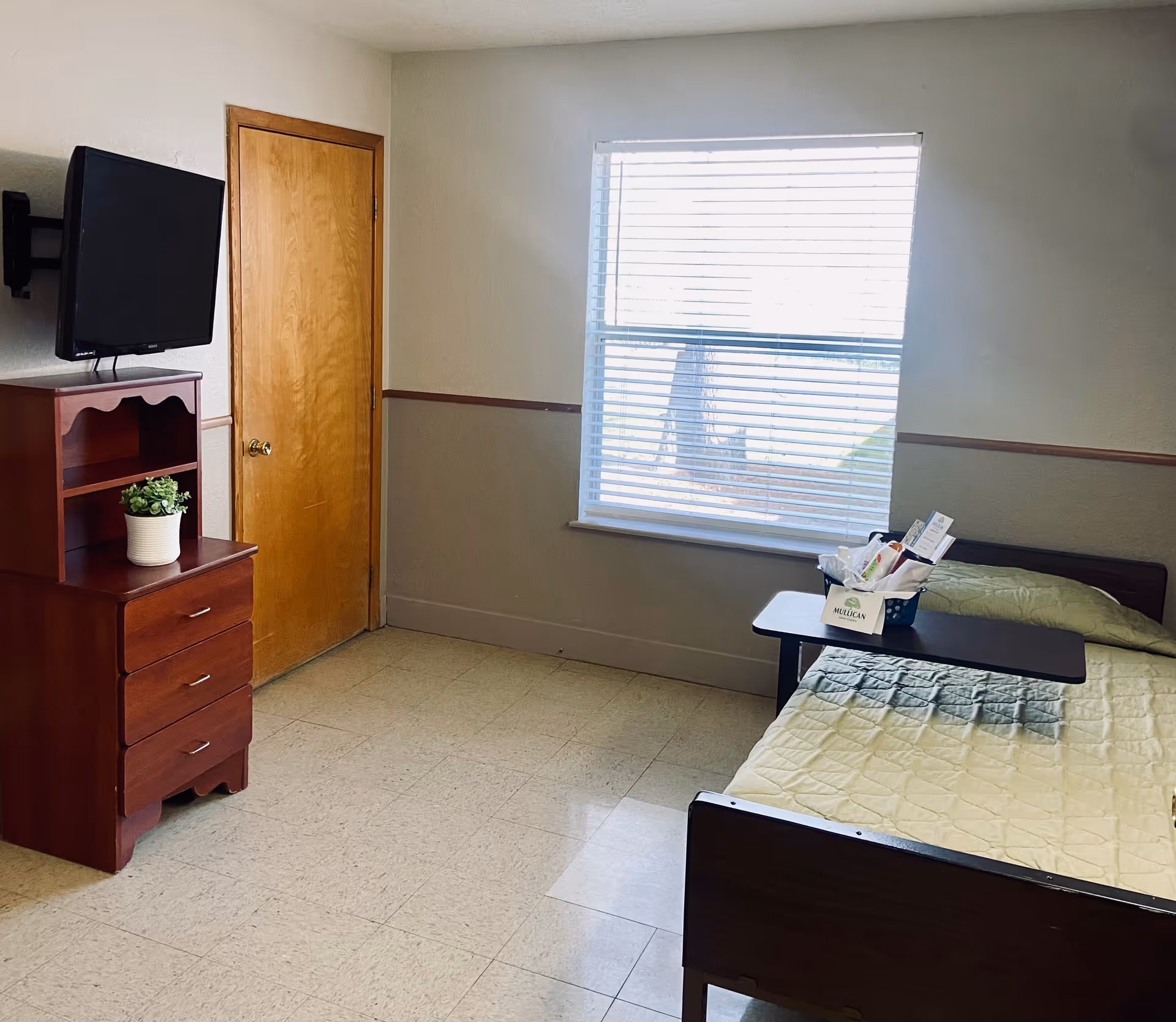 A simple room with a single bed covered with a light green quilt, a small table with a basket of items on it, a wooden dresser with a small potted plant, a wall-mounted TV, a closed wooden door, and a window with blinds letting in natural light.