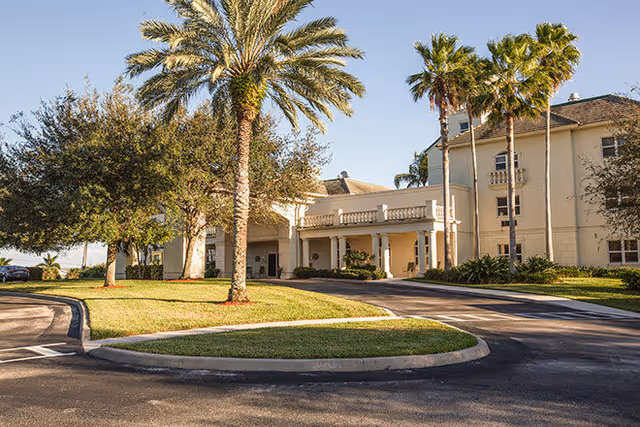 Exterior view of a senior living facility building with a driveway, palm trees, and well-maintained landscaping under a clear blue sky.