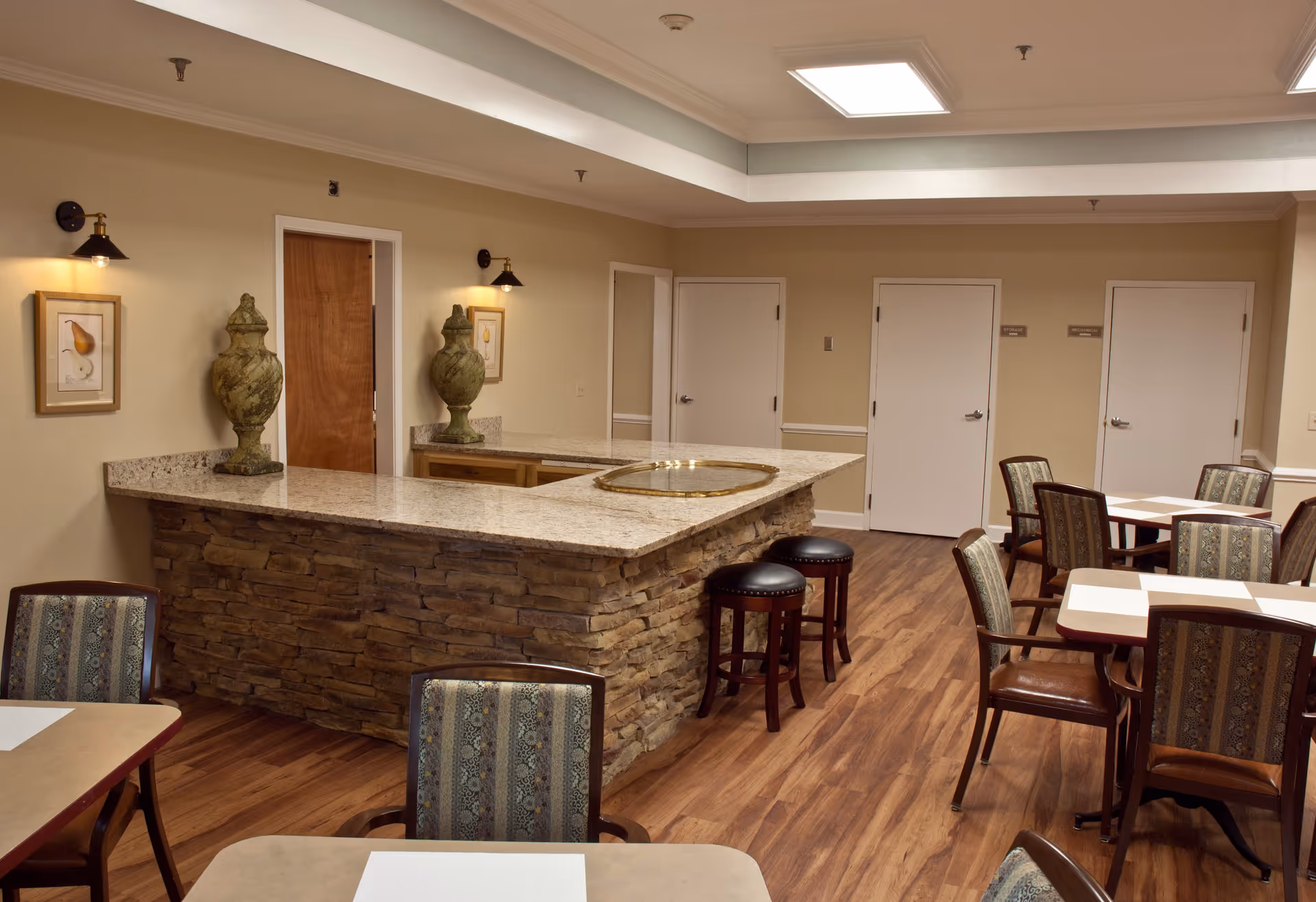 Interior view of a senior living community dining area featuring a stone and granite countertop bar with two decorative urns and a brass tray. The room has wooden floors, several tables with patterned upholstered chairs, beige walls with framed artwork, and three white doors labeled storage and mechanical.