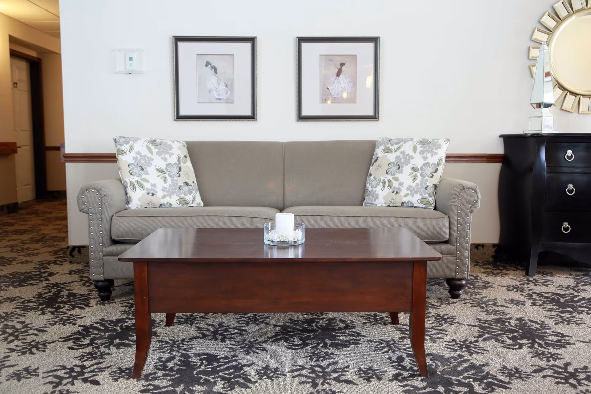 A cozy living room area featuring a beige sofa with two floral patterned cushions, a dark wooden coffee table with a candle centerpiece, two framed artworks on the wall above the sofa, a black chest of drawers with silver handles, and a decorative round mirror on the wall. The floor is carpeted with a floral pattern.