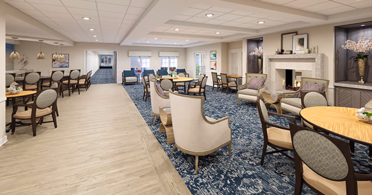 A spacious and well-lit common area in a senior living facility featuring multiple seating arrangements including round tables with chairs, armchairs, and a fireplace with decorative vases and artwork. The room has a mix of wood flooring and patterned carpet, with large windows allowing natural light to enter.