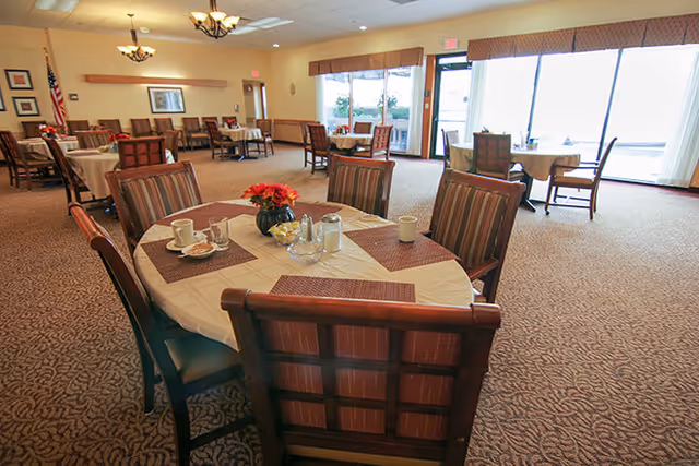 A spacious dining room with round tables covered in white tablecloths and brown placemats. Each table is surrounded by wooden chairs with striped cushions. One table in the foreground has a small flower arrangement, salt and pepper shakers, and cups. Large windows and glass doors allow natural light to fill the room. The room has beige walls, carpeted floors, and ceiling light fixtures.