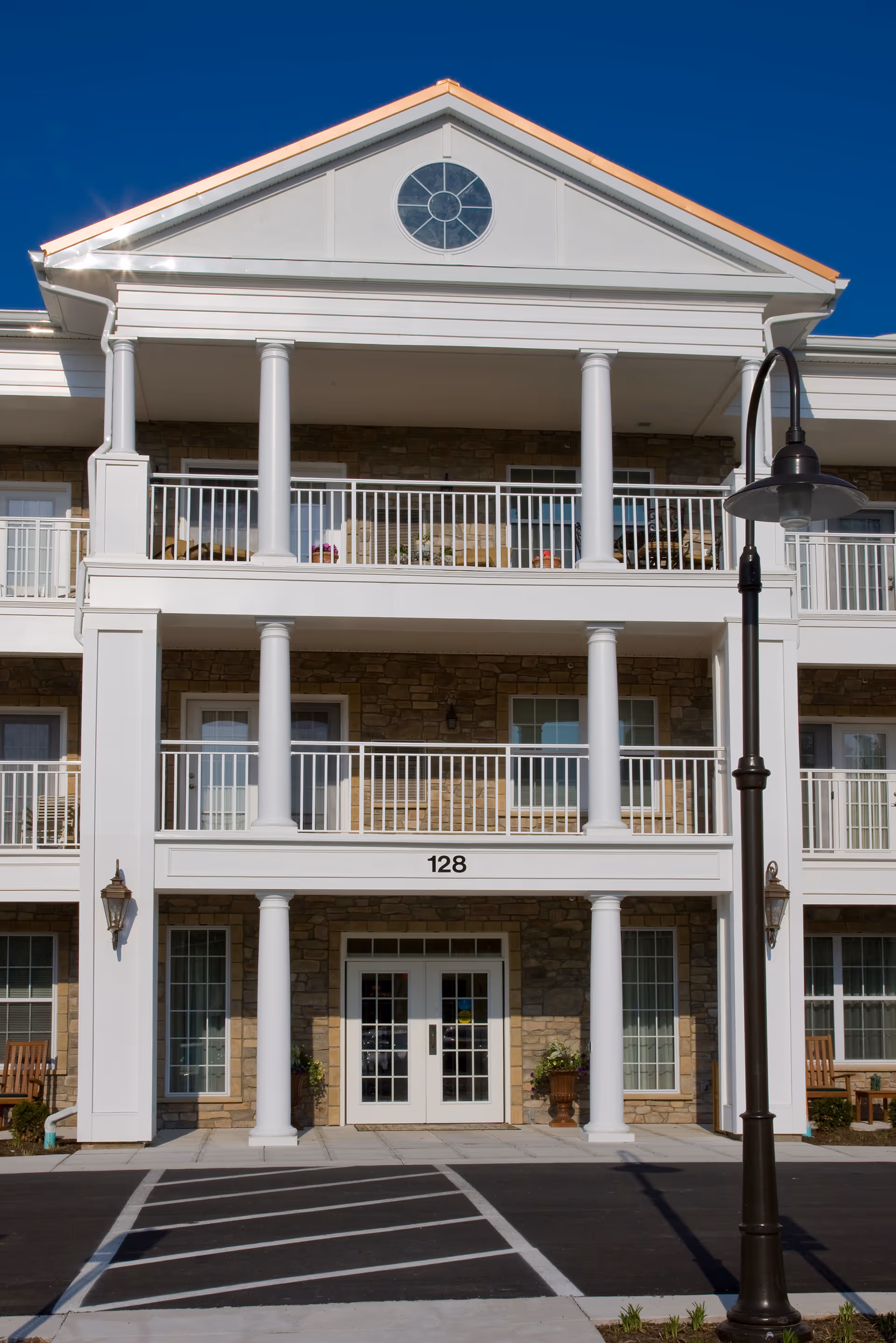 Front exterior view of a senior living facility building with white columns, balconies, and a stone facade under a clear blue sky. The building number 128 is displayed above the entrance, which has double glass doors. A street lamp is visible on the right side of the image.