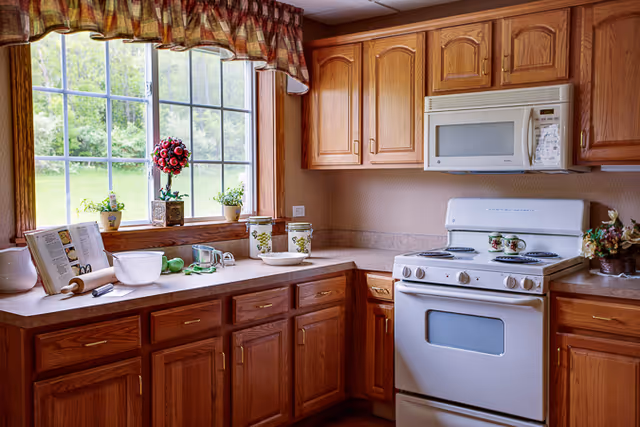 A cozy kitchen with wooden cabinets and a white stove and microwave. The countertop holds various kitchen items including a rolling pin, mixing bowl, measuring cups, and decorative canisters. A large window with a plaid valance lets in natural light and shows a green outdoor view. Small potted plants and a flower arrangement decorate the windowsill.