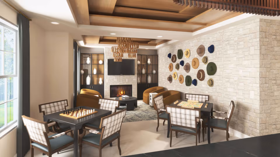 A cozy common area with two tables set up for board games, including chess and Scrabble. The room features comfortable armchairs, a fireplace with a TV mounted above it, built-in shelves with decorative vases, and a wall decorated with various hats. Large windows allow natural light to brighten the space.