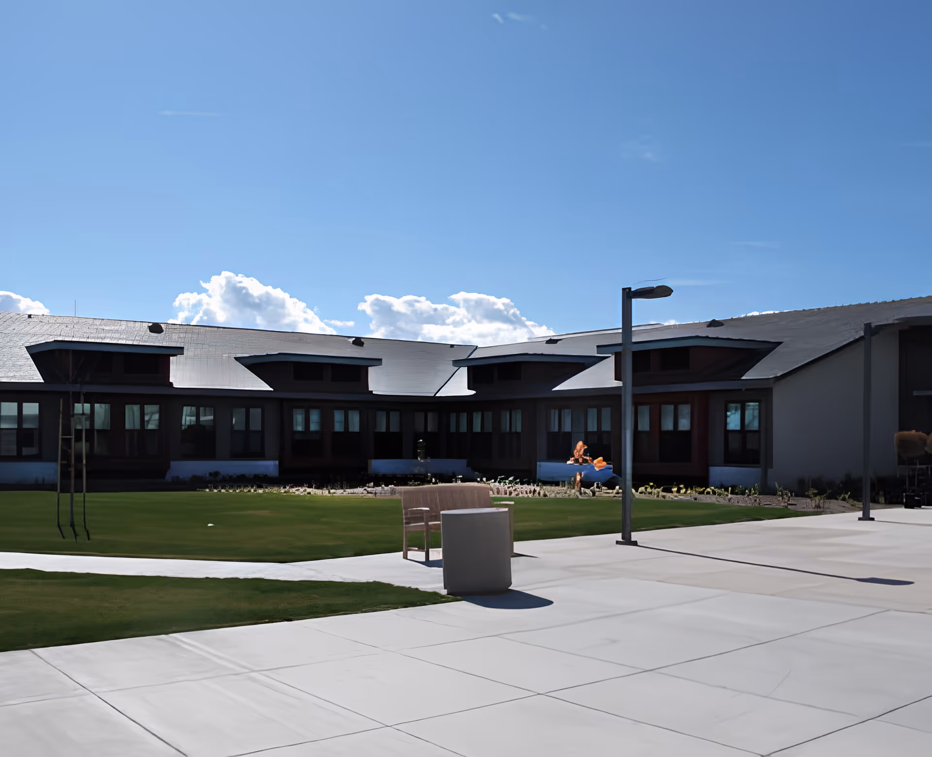 Exterior courtyard of a modern senior living building with a paved patio, lawn, benches, lamp posts and a low roofline under a blue sky.