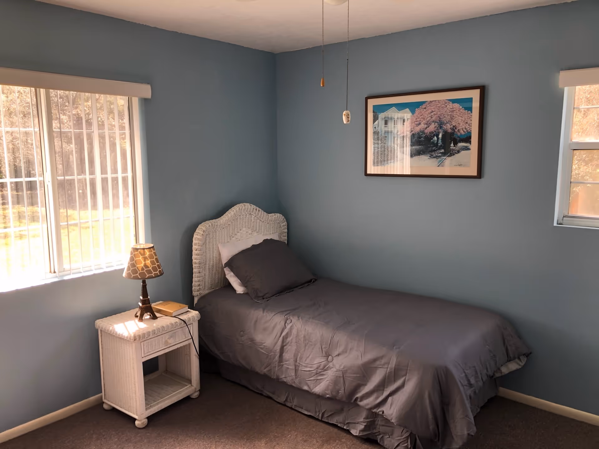 A small bedroom with light blue walls, a single bed with gray bedding and a white wicker headboard, a white wicker nightstand with a lamp and a book, two windows with vertical blinds, and a framed picture of a house and a pink tree on the wall.