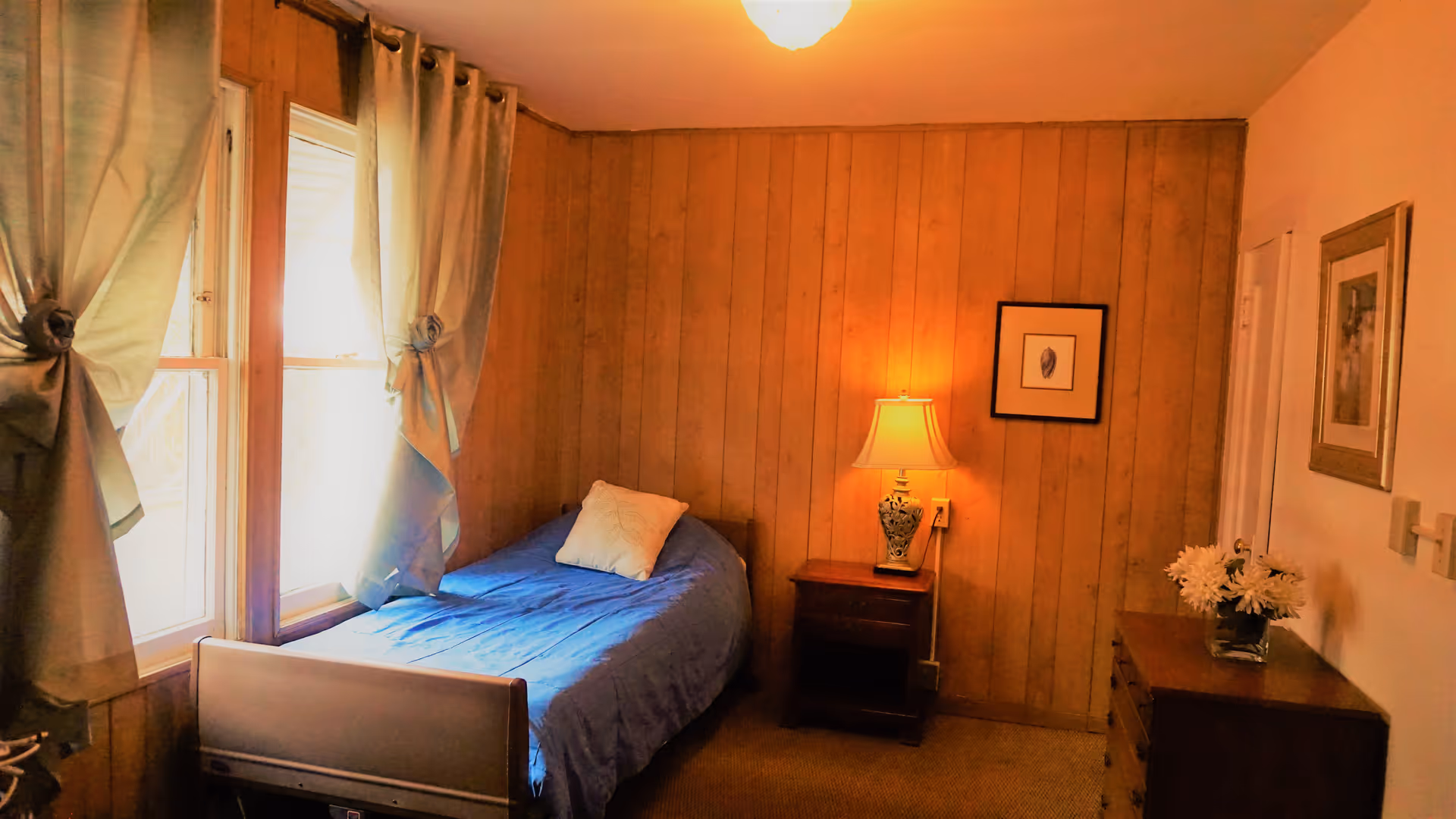 A small bedroom with wood-paneled walls, a single bed with a blue bedspread and a white pillow, a nightstand with a decorative lamp, a dresser with a vase of white flowers, two framed pictures on the walls, and two windows with tied-back curtains letting in natural light.