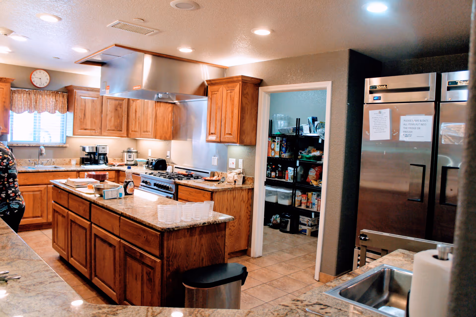 Large communal kitchen with a central island, wooden cabinets, stainless-steel appliances and an open pantry stocked with supplies.