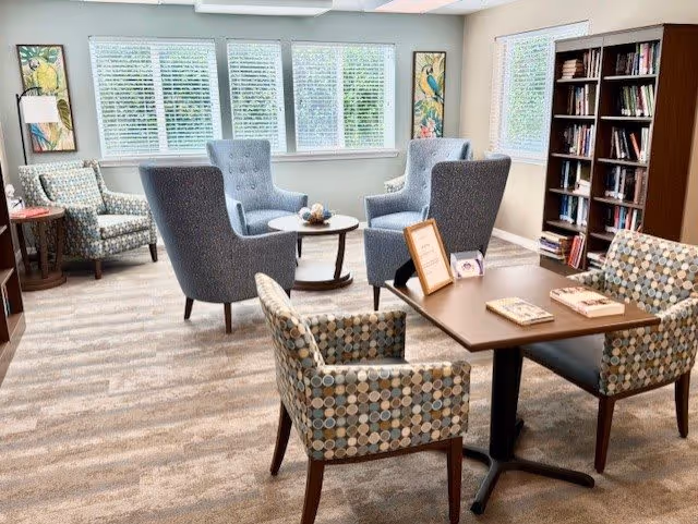 A bright and cozy common area with large windows covered by white blinds. The room features a seating arrangement with four blue armchairs around a small round table, two patterned armchairs at a rectangular table with books and a framed sign, and a bookshelf filled with books. The walls have framed artwork with colorful birds.