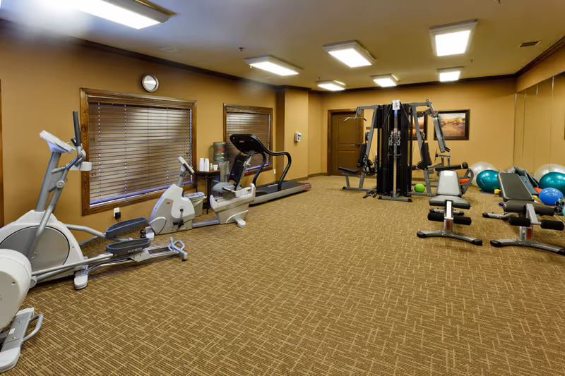 Interior view of a fitness room with exercise equipment including stationary bikes, a treadmill, weight machines, benches, and exercise balls. The room has carpeted flooring, beige walls, two windows with wooden blinds, a clock, and bright ceiling lights.