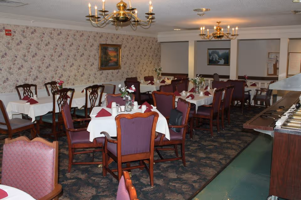 A dining room with multiple tables covered with white tablecloths and set with maroon napkins and small flower arrangements. The room has floral wallpaper, carpeted floor, and brass chandeliers hanging from the ceiling. There is a buffet counter on the right side and framed paintings on the walls.