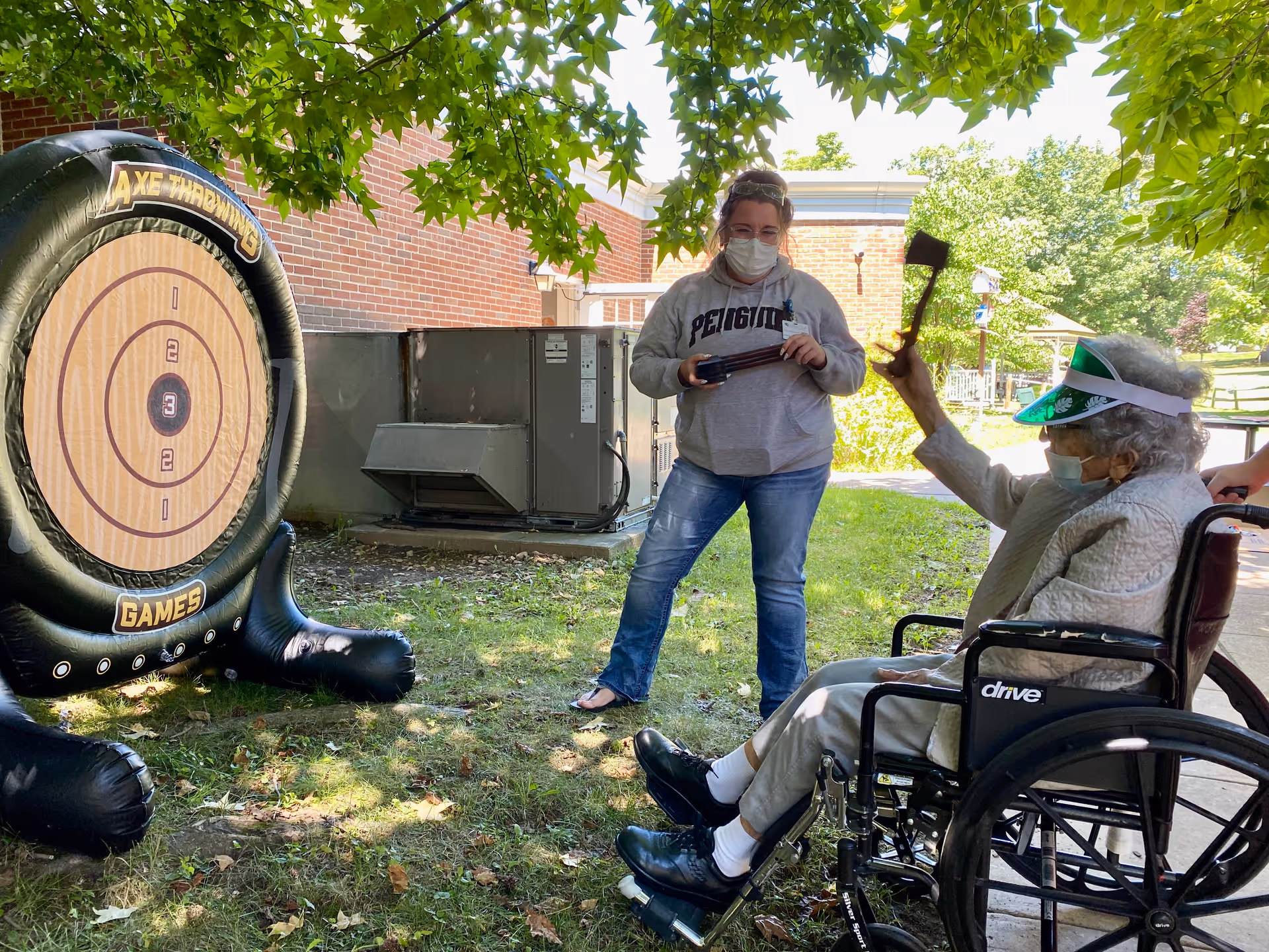 An elderly person in a wheelchair wearing a visor and face mask is playing an outdoor axe throwing game, holding an axe in the air. A woman wearing a face mask and a grey sweatshirt stands nearby, holding several axes. The scene is set outside near a brick building with green trees providing shade.