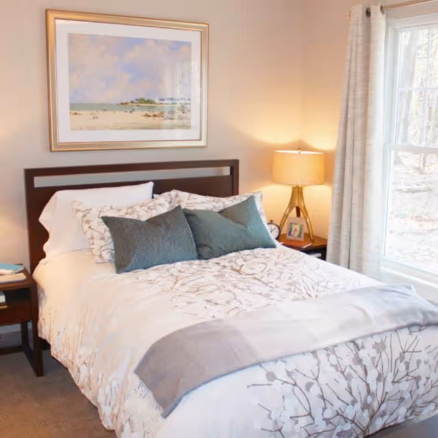A cozy bedroom with a neatly made bed featuring white and patterned bedding, two teal accent pillows, a wooden headboard, a nightstand with a lamp, a small clock, and a framed photo. A large window with light curtains allows natural light to brighten the room. A framed beach-themed painting hangs on the wall above the bed.