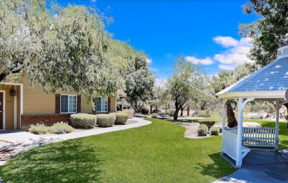 Well-maintained outdoor courtyard with green lawn, a white gazebo, and a single-story building under a blue sky.