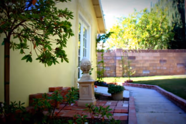 Outdoor pathway beside a yellow building with a window, bordered by a brick planter with green plants and a decorative stone sculpture. A brick wall and trees are visible in the background under a bright sky.