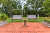 Two metal-framed outdoor swing benches with beige cushions facing each other on a red brick patio, surrounded by green grass, trees, and raised garden beds with plants on either side.