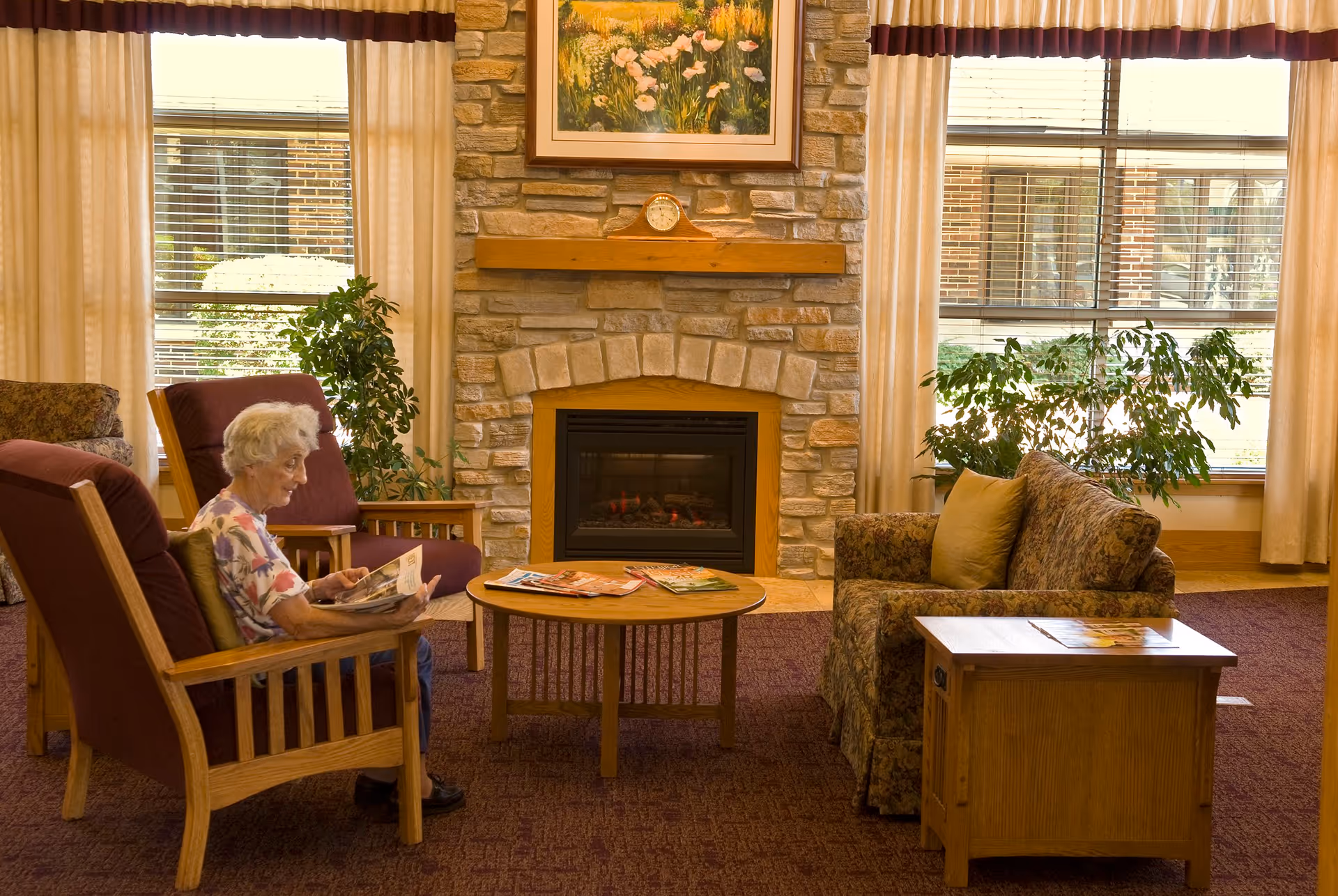 An elderly woman sitting in a wooden armchair with maroon cushions, reading a magazine in a cozy living room. The room features a stone fireplace with a wooden mantle, a round wooden coffee table with magazines, a floral-patterned sofa with a green pillow, large windows with cream-colored curtains, and green plants near the windows.