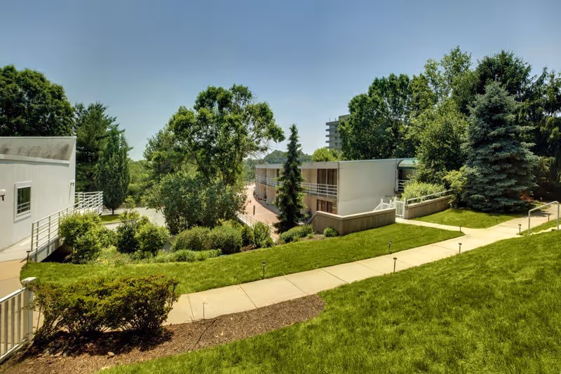 Outdoor view of a senior living facility with well-maintained green lawns, bushes, and trees. There are paved walkways and modern white buildings in the background under a clear blue sky.