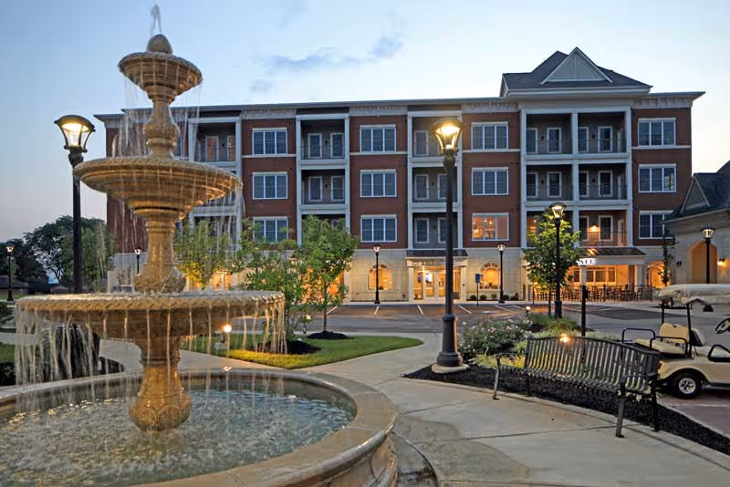 Outdoor view of a senior living community building at dusk with a multi-tiered water fountain in the foreground, lamp posts, benches, and a golf cart parked on the right side.