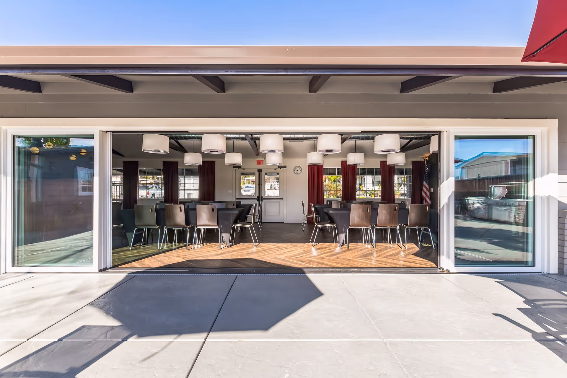 Open sliding glass doors reveal a community meeting room with tables, chairs, and hanging pendant lights viewed from the outdoor patio.