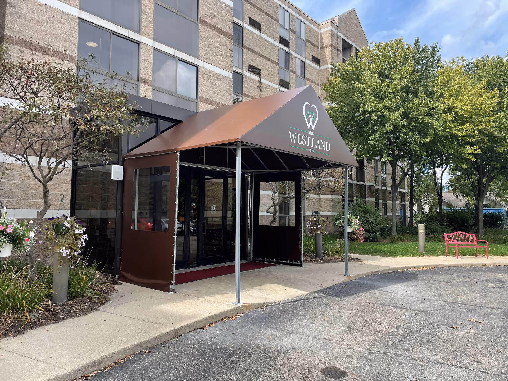 Entrance to The Westland House facility with a brown canopy over the doorway, a beige brick building with multiple windows, surrounding greenery including trees and flower pots, and a red bench on the right side.