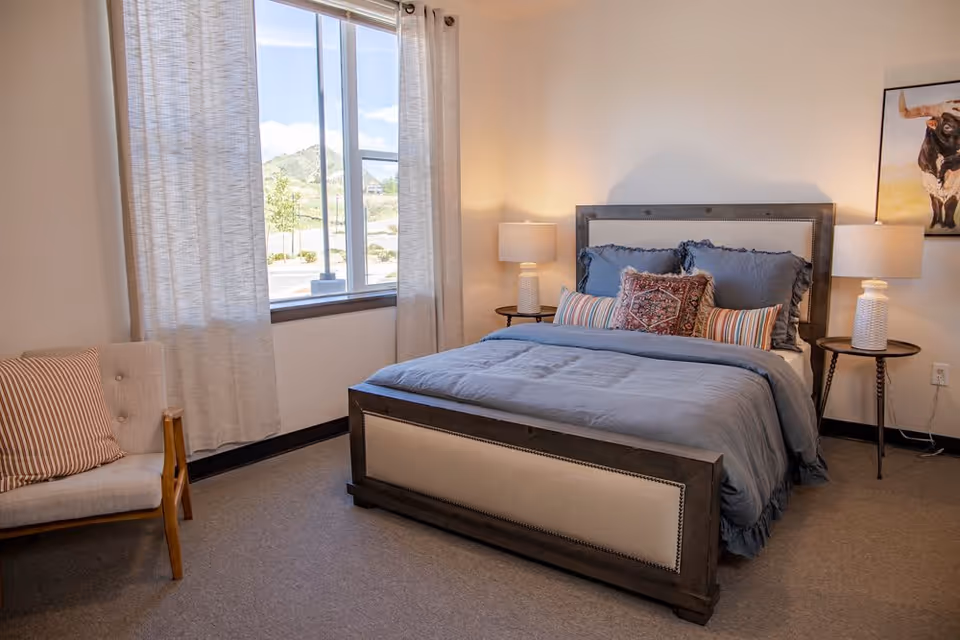 A cozy bedroom with a large bed featuring blue bedding and multiple decorative pillows. There are two bedside tables with matching lamps on each side of the bed. A window with light curtains lets in natural light, and a cushioned chair with a striped pillow is positioned near the window. A framed picture of a bull hangs on the wall to the right.